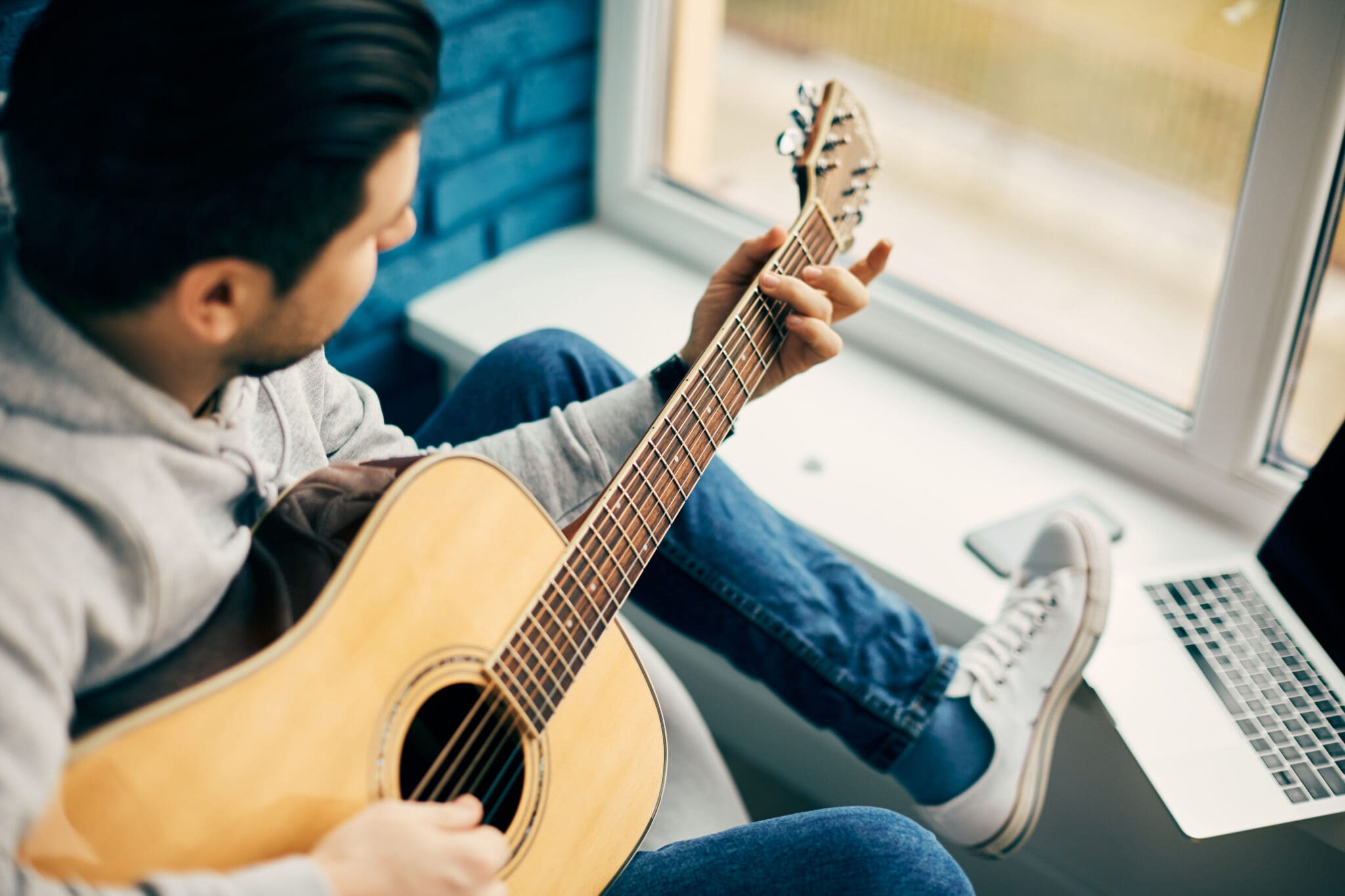 close-up of guitarist holding guitar and sitting near window with laptop on the windowsill